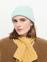 A person wearing the Cashmere Beanie in light blue, gold hoop earrings, a brown sweater, and a yellow soft knit scarf looks at the camera against a plain white background.