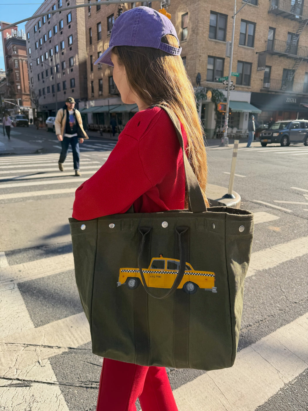 A woman in a purple cap and red outfit crosses a city street, carrying the Limited Edition Painted Perfect Weekday Taxi Tote—its green, hand-painted NYC taxi design glowing in the sunlight, reflecting Brooklyn’s creative spirit.