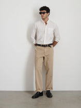 A man stands with his hands in his pockets against a plain white wall and gray floor, wearing sunglasses, beige pants, black shoes, and the Mill Shirt in Cotton Poplin featuring French seams and real shell buttons.