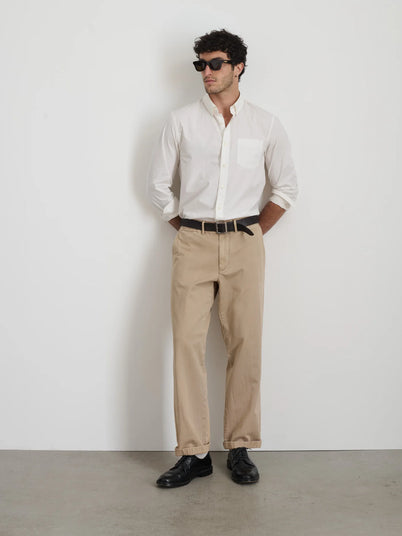 A man stands with his hands in his pockets against a plain white wall and gray floor, wearing sunglasses, beige pants, black shoes, and the Mill Shirt in Cotton Poplin featuring French seams and real shell buttons.