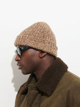 A person in profile view wears the Cashmere Marled Beanie in tan, along with dark sunglasses and a brown jacket with a shearling collar, standing against a plain white background.