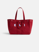 The Painted Perfect Weekend Tote is a large red canvas bag with two handles, featuring "ELI" in bold white and blue letters on the front, displayed on a plain white background.