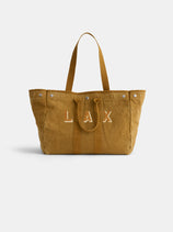 The Painted Perfect Weekend Tote is a large tan canvas bag with two handles, silver rivets, and "LAX" stitched in white on the front, displayed against a plain white background.