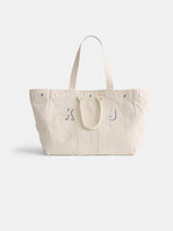 The Perfect Weekend Tote is a spacious off-white cotton bag with two handles and silver rivets. Featuring gray "K" and "J" letters on the front, it's ideal for fans of personalized style. The background is plain and light-colored.