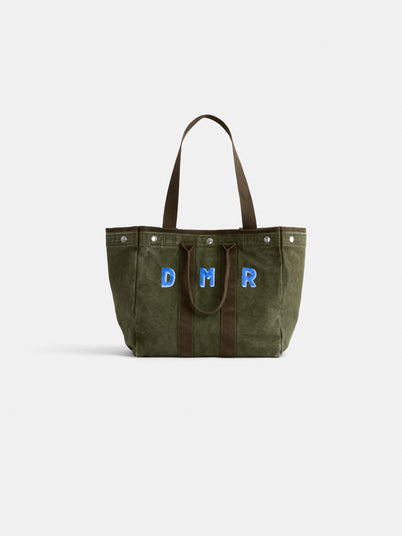 The Perfect Weekday Tote is a green suede bag with brown handles, featuring large blue and white "DMR" letters monogrammed on the front, displayed against a plain white background.