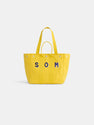 The Painted Perfect Weekday Tote is a bright yellow canvas bag with two handles and blue "SOM" letters outlined in white on the front, shown against a plain white background.