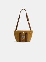 The Painted Mini is a brown canvas shoulder bag with two short handles, an adjustable strap, and a front pocket decorated with a blue and white oval emblem, shown against a plain white background.