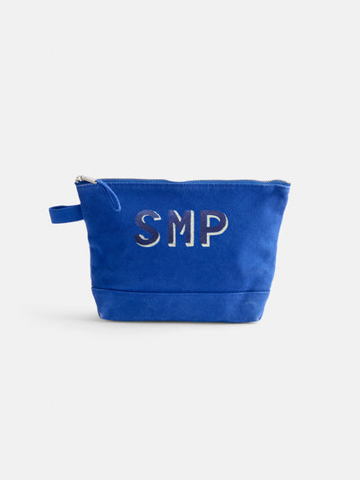 The Perfect Canvas Pouch is a blue cotton makeup bag with a zippered closure, small side handle, and silver SMP letters on the front. Its vintage military style is showcased against a plain white background.