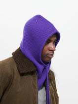 A person wearing the Balaclava in Cashmere—a purple knit hood—paired with a brown jacket featuring a shearling collar, looking to the side against a plain white background.
