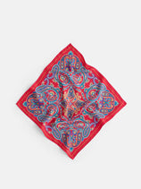 The Paisley Scarf In Silk, crafted in Italy, is a square silk scarf featuring a vibrant red border and an intricate paisley and floral pattern in blue, orange, and yellow tones, displayed on a light background.