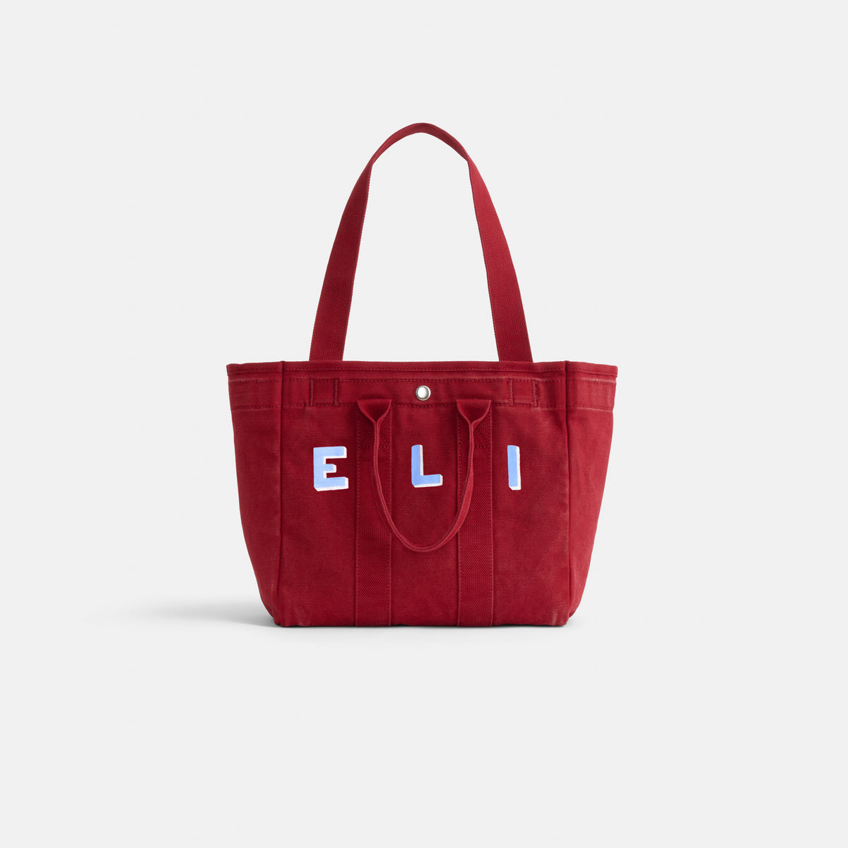 The Painted Perfect Daily Tote is a red bag with two handles, a front pocket, and "ELI" in white applique on the front. The background is plain white.