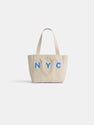 The Painted Perfect Daily Tote is a beige canvas bag with two handles and blue "NYC" lettering on the front, shown on a plain white background.