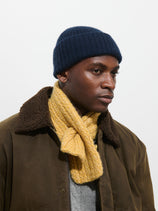A person wearing a dark blue knit beanie, a brown jacket with a shearling collar, the Bedford Scarf in Cashmere in yellow, and a grey sweater, looking slightly to the side against a plain background.