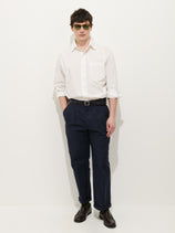 A man stands against a white background wearing dark sunglasses, a white button-up shirt with rolled sleeves, Double Pleated Pant In Vintage Twill in navy blue, a black belt, and dark brown dress shoes, hands in his pockets.