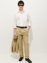 A man in sunglasses, a white button-down with rolled sleeves, Double Pleated Pant In Vintage Twill, and dark dress shoes stands against a plain background, holding a matching beige jacket in his left hand.