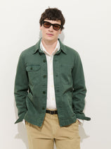 A man wearing sunglasses, a Garment Dyed Work Jacket In Recycled Denim over a white shirt, and beige pants stands against a plain white background with his hands in his pockets.