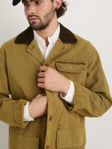 A man fastens the front buttons of the Frontier Jacket In Canvas, featuring a tan color and corduroy collar over a white shirt. His face is partially visible, and he also wears a navy cap.
