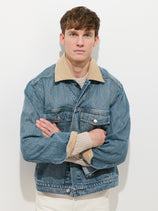 A person with short brown hair stands in front of a white background, arms crossed, wearing the unisex Denim Jacket with a corduroy collar over a beige sweater.