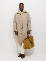 A man in the Highgate Trench In Double-Faced Cotton, cream pants, a light yellow sweater, and brown shoes stands against a white background, holding a large mustard suede bag in his left hand.