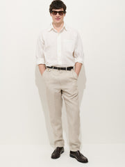 A man stands against a white background wearing sunglasses, a white rolled-sleeve shirt, the Standard Pleated Pant in flax linen, a black belt, and dark brown dress shoes. His hands are in his pockets.