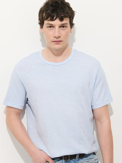 A young man with short dark hair stands against a plain white background, wearing jeans and the Standard T-Shirt In Slub Cotton, a light blue men’s tee made from breathable fabric, looking directly at the camera with a neutral expression.