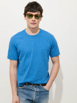 A man wearing the Standard T-Shirt In Slub Cotton in bright blue, paired with blue jeans and green-tinted sunglasses, stands against a plain white background with one hand in his pocket.
