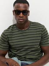 A man in dark sunglasses and the Standard Striped Tee In Cotton, featuring green stripes, sits against a white background, looking straight at the camera.