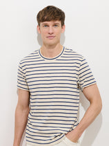 A young man with short brown hair wears the Standard Striped Tee In Cotton, featuring navy blue horizontal stripes and crafted from breathable slub cotton. He stands against a plain white background, looking at the camera with a slight smile.