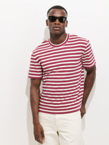 A man in dark sunglasses and light-colored pants stands confidently against a white background, wearing the Crosby Striped Tee In Heavy Slub Cotton—a red and white striped, easy fit short-sleeve shirt.