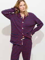 A woman poses against a plain white background in The Striped Waffle Sleeping Shirt, featuring blue and red stripes, white buttons, and 100% cotton fabric; one hand behind her head and the other on her hip.