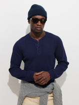 A man stands against a plain white background wearing dark sunglasses, a navy beanie, light pants with a gray sweater tied around his waist, and the Austin Henley In Thermal Cotton—a navy lightweight breathable shirt.