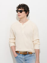 Wearing the Austin Henley In Thermal Cotton, a person stands against a plain white background with one hand in their pocket. They pair the cream-colored lightweight, breathable henley shirt with black sunglasses, light blue jeans, and a brown belt.