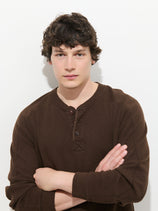 A young man with curly brown hair, wearing the Austin Henley In Thermal Cotton in a brown pigment-dyed finish, stands against a white background with his arms crossed and a neutral expression.