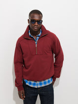 A man wearing sunglasses, the Noah Half Zip in Cotton Fleece in maroon, and a blue plaid shirt stands against a plain white background with one hand in his pocket.