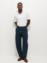 A man poses confidently against a white background, wearing a white polo shirt, brown shoes, black belt, and the AM Arc 5 Pocket Jean in Turkish dark blue denim with an arc-shaped leg. His hands are behind his back as he looks at the camera.