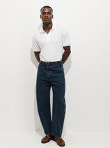 A man faces the camera against a white background, wearing a white polo shirt, black belt, brown lace-up shoes, and AM Arc 5 Pocket Jeans—high-waisted blue jeans with a Hollywood waist and tapered fit. His hands are behind his back.