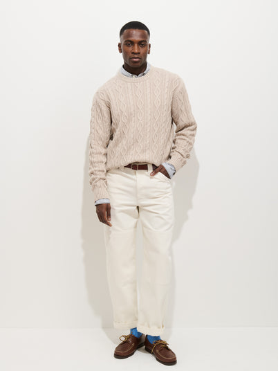 A man stands against a white background wearing the Aaron Cable Sweater in Cotton, styled over a collared shirt with white pants, a brown belt, and brown loafers. With his left hand in his pocket, he gazes confidently at the camera.