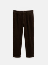 The Standard Pleated Pant In Cord, a dark brown 100% cotton corduroy pant made in NYC, features front pleats, rolled cuffs, and a button-zip closure. Shown laid flat on a white background.