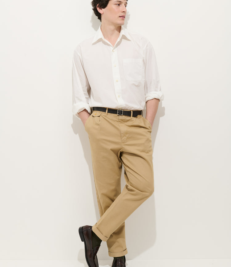 A person wearing a white long-sleeve button-up shirt, Standard Pleated Pant in Chino, and dark shoes stands against a plain white background with hands in pockets and one leg crossed over the other.