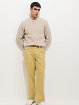 A man stands against a white wall wearing a beige cable-knit sweater, light yellow Field Pant In Herringbone with a front pocket, and white sneakers. His hands are in his pockets as he gazes slightly to the side.