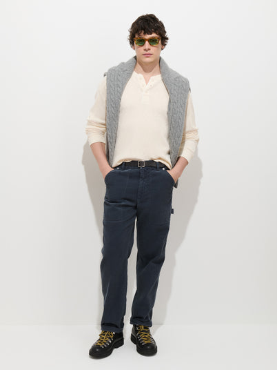 A person stands against a white background with hands in pockets, wearing sunglasses, a cream long-sleeve shirt with a gray sweater draped over their shoulders, black shoes, and the Painter Pant in Fine Wale Corduroy in a men’s straight fit and vintage wash.