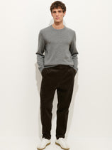 A man stands against a plain white background wearing a light grey sweater, white sneakers, and the Standard Pleated Pant In Cord—dark brown corduroy pants made in NYC—with his hands in his pockets.