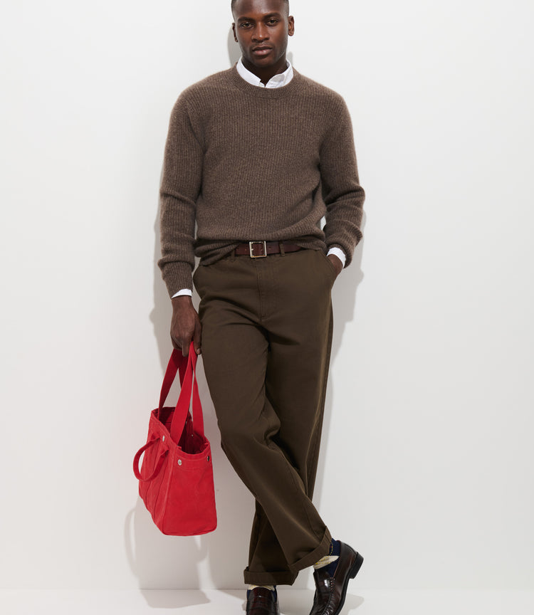 A man in a brown sweater, Flat Front Chino in Vintage Wash men’s cotton pants, and dark shoes stands against a white wall, holding a bright red tote bag in his left hand. His right hand is in his pocket, and he looks confidently at the camera.