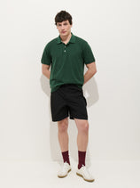 A man stands against a white background, wearing a dark green polo shirt and Camino Short in Crinkly Nylon. He completes his look with burgundy socks and white sneakers, hands behind his back, looking slightly to the side with a soft smile.