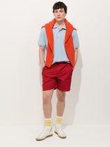 A person stands against a white background wearing the Camino Short in Crinkly Nylon, a light blue polo shirt, an orange sweater over their shoulders, white socks with yellow stripes, and white sneakers. Their hands are in their pockets.
