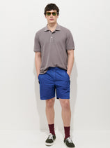 A man wears sunglasses, a gray polo shirt, Camino Short in Crinkly Nylon (vintage-inspired bright blue), maroon socks, and black sneakers, standing against a plain white background with his hands in his pockets.