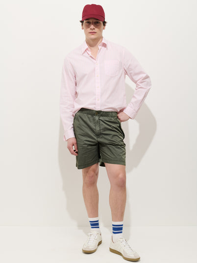 A person wearing a red cap, the Mill Shirt In Thin Stripe (light pink, 100% cotton button-down), green shorts, white socks with blue stripes, and white sneakers stands against a plain white background.