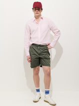 Against a white background, a person wears a red cap, light pink button-up shirt, Camino Short In Crinkly Nylon, white sneakers, and white socks with blue stripes. One hand is in their shorts pocket as they look at the camera.