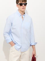 A man in dark sunglasses wears the Mill Shirt In Cotton Poplin, a light blue 100% cotton button-down, paired with white pants against a plain light background, with one hand in his pocket and the other holding a red item out of frame.