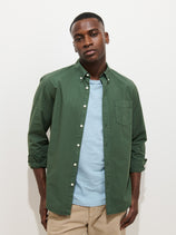 A man wears the Mill Shirt In Cotton Poplin, styled in green over a light blue T-shirt with beige pants, standing against a plain white background and looking at the camera with a neutral expression.